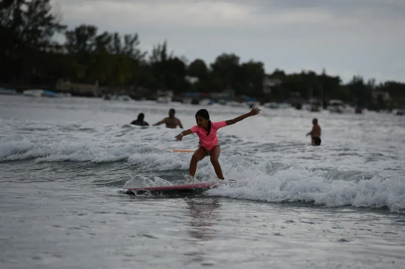 A Duck in a pink rashie rides a wave in front of the beach, arms out.